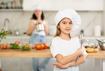 Little girl in chefs hat and white T-shirt stands in kitchen near table, her mother in background.