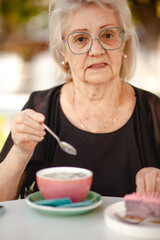 A woman aged 75-80 sit in the cafe of the cityand drink tea or coffee wit dessert. An elderly madam is wearing jeans and a  shirt