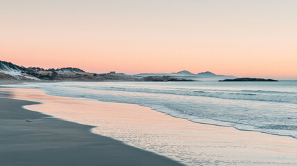 Obraz premium An unexpected winter beach scene with frosty sand, gentle waves rolling in, and a morning sky tinted with pale pink and orange. Room for text in the sky