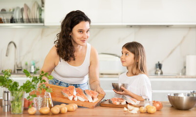 Cheerful middle-aged woman holding wooden board with fish pieces standing at kitchen-table with little daughter