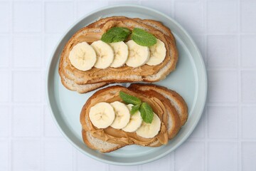 Tasty sandwiches with peanut butter, banana and mint on white tiled table, top view