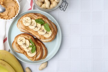 Tasty sandwiches with peanut butter, banana, chia seeds, nuts and mint on white tiled table, flat lay. Space for text