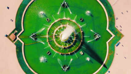 Aerial View of Circular Green Fountain with Jets. Aerial shot of a large circular fountain with symmetrical water jets and green water, set in an open plaza with visible pathways. © Iryna