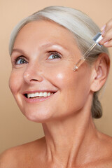Beauty portrait of a mature woman with gray hair applying facial skin care serum with a dropper, highlighting her skincare routine on a beige background...
