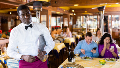 Portrait of sad African-american man waiter with tray in restaurant.