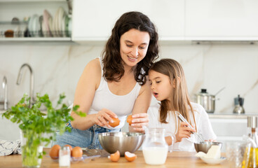 Mom teaches her daughter how to make dough, breaks eggs into bowl. Woman explains to her daughter successful recipe and rules for making dough for dessert. Homemade food and little helper.