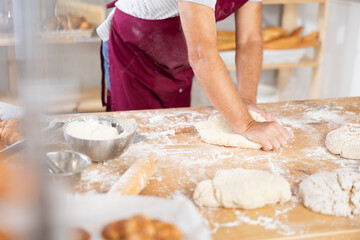 Close-up of professional male baker hands in dark red apron kneading dough on flour-dusted wooden table in artisan bakery, highlighting craftsmanship of traditional breadmaking