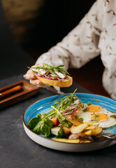 Hand holding sardine toast over a vibrant breakfast plate with eggs, salad, and potatoes