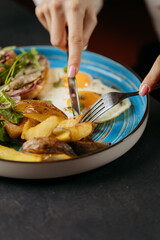 Close-up of hands cutting into a breakfast plate with roasted potatoes, eggs, and greens