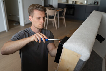 A young man in a gray T-shirt is assembling a sofa by himself at home.