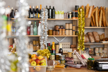 Variety of goods on counter, vacuumed meat delicacies and cheeses, fresh apples. Shopping hall of grocery minimarket is decorated with tinsel for New Year. In background there are shelves with bread