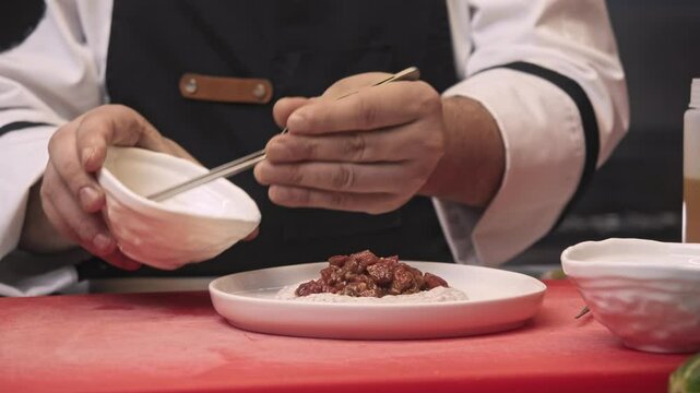 Cook uses tongs to add seeds of Dijon mustard to steak tartare, which is what decorates exquisite dish of French cuisine from raw beef. Close up