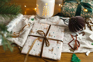 Merry Christmas! Stylish christmas gift, modern paper ornaments and pine branches on rustic table with golden christmas lights. Atmospheric christmas eve. Modern wrapped present