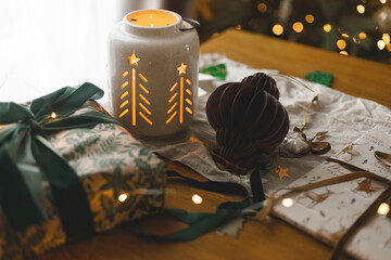 Merry Christmas and Happy holidays! Stylish christmas gift, glowing lantern, modern paper ornaments on table against golden christmas tree lights. Atmospheric christmas eve, holiday magical time