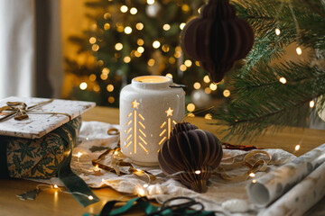 Merry Christmas and Happy holidays! Stylish christmas gift, glowing lantern, modern paper ornaments on table against golden christmas tree lights. Atmospheric christmas eve, holiday magical time