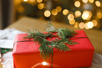 Merry Christmas and Happy holidays! Stylish christmas red gift with cedar wreath on table against golden christmas tree lights. Atmospheric eve, holiday magical banner