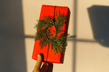 Hand holding stylish christmas red gift with modern cedar wreath in sunlight on background of grey wall. Merry Christmas! Atmospheric christmas time, holiday banner