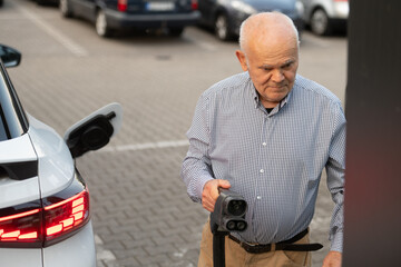 Senior Man Holding EV Charging Plug Near Electric Car in Parking Lot