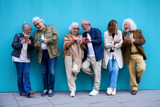 Group of six elderly Caucasian happy people leaning on blue wall using mobile phone. Cheerful mature grey hair couples friends together having fun enjoying looking smiling cellular telephone outdoors