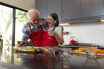 Senior couple cooking together in kitchen, enjoying wine and using tablet, at home