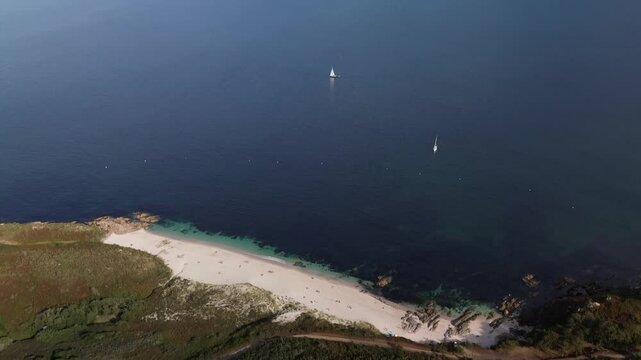 `Pan up from Melide Beach. White sand and turquoise waters surrounded by lush dunes and vegetation. Two sailboats moored on calm sea under bright sun. Pontevedra Estuary and Ald&aacute;n Bay in background.