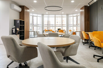 Empty working area in an open office. White round table surrounded by chairs in grey and yellow colors. Big window background