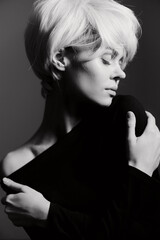 Elegant woman with short blonde hair wearing a black off-shoulder top, softly embracing herself in a monochrome setting. Artistic expression and beauty concept.