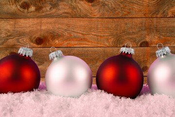 Four Christmas ornaments arranged in a row, with alternating colors of red and silver, placed on a bed of artificial snow. Wooden plank in background with copy space for your text.