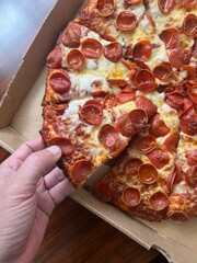 Hand taking a piece of cheesy large pepperoni pizza with cheese from a cardboard box. Takeout delivery to-go from a restaurant.