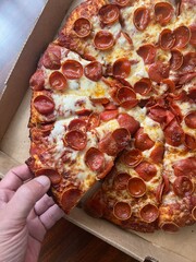 Hand taking a piece of cheesy large pepperoni pizza with cheese from a cardboard box. Takeout delivery to-go from a restaurant.