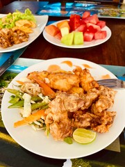 Fried chicken and fried potatoes with a vegetable garnish of carrots, peas, cabbage, sprouts, with a lime  wedge on a white plate. In the background is a fruit plate with watermelons, melons, papaya.