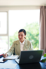 A professional woman in a light suit sits at a desk, focused on her work with documents and a laptop in front of her. The bright office setting