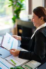 A businesswoman in a black blazer attentively reviews business documents in her office. She sits at her desk in a modern workspace, focused on reading the papers