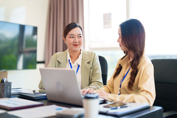 Obraz premium Two businesswomen in formal attire collaborate on a laptop in a bright office. They are both wearing blue lanyards and discussing ideas in a modern workspace.