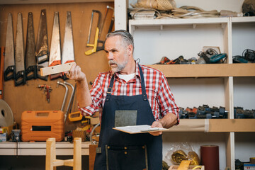 Carpenter in a plaid shirt and apron inspecting a piece of wood while holding a clipboard in...