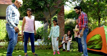 Indian Asian multigenerational family enjoying outdoor camping time as grandpa and son play football with granddaughters, while grandma and mother watch and cheer with smiles - Powered by Adobe