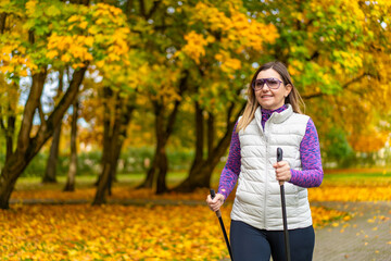 Portrait of beautiful middle aged woman in sports wears practicing nordic walking on alley full of yellow fallen leaves in park on autumn day. Front view. Autumnal training of nordic walking