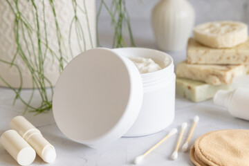 Opened cream jar with white blank lid on marble near green plant close up, cosmetic mockup