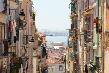 A view to the sea from a colourful, narrow and chaotic street in the old neighbourhood of Alfama, Lisbon, Portugal. Historic buildings with clotheslines, plants and antennas create a beautiful scene.