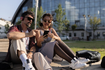 Young couple enjoying music together outdoors on a sunny day, with headphones and mobile device, sitting on wooden bench in urban park setting
