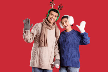 Fototapeta premium Happy young couple in winter clothes waving hands on red background