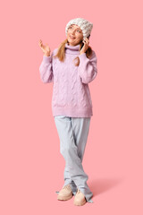 Beautiful mature woman in stylish lilac sweater and knitted hat talking by mobile phone on pink background
