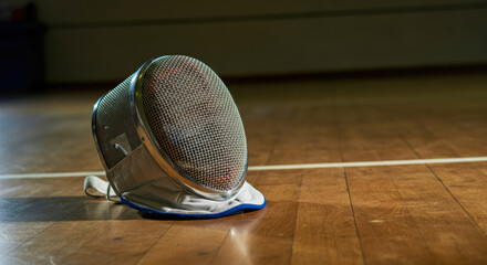 A fencing mask rests on the wooden floor of a gym after an intense match during an afternoon practice session