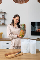 A cheerful young woman in loungewear holding a fresh pineapple in her kitchen. Promoting healthy...