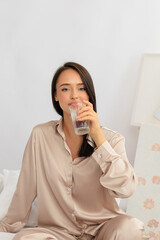 A young woman in cozy loungewear enjoys a refreshing glass of water while relaxing at home. Promoting hydration, wellness, and self-care in a serene indoor setting.