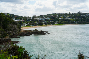 Obraz premium Scenic Coastal View Featuring Calm Waters, Rocky Outcrops, and Lush Greenery with Residential Dwellings on the Hillside near the Beach