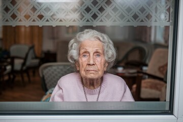 A thoughtful elderly woman with gray hair, looking out of a window in a nursing home. Her expression is contemplative, suggesting a mix of longing and quiet reflection. 