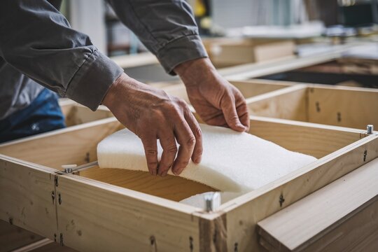 Close-up view of a skilled artisan meticulously installing foam padding into a wooden frame for upholstered furniture. 