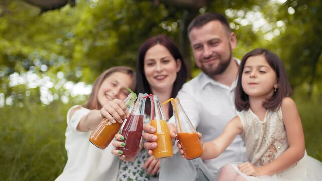 Happy young family with two children drinking fresh juice while sitting on blanket at green garden. Caucasian parents with two cute daughters drinking juice in glass bottle from colorful straws.