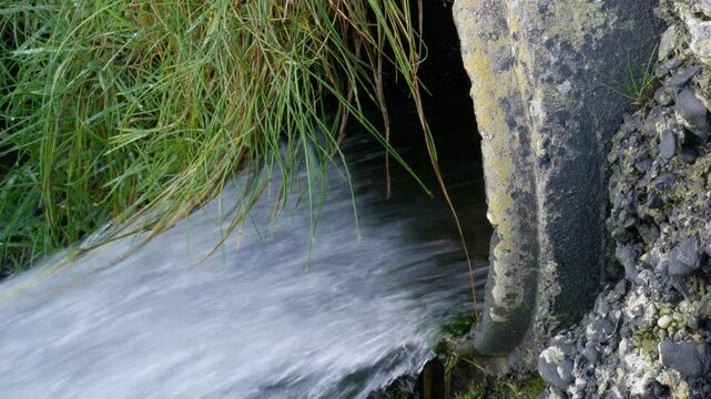 A concrete culvert with a smooth, dark surface has water flowing out of it. The water appears to be clear and moving quickly out of the pipe.